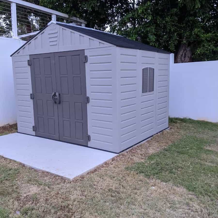 Gray outdoor shed with dark double doors and a small window, situated on a concrete slab in a fenced grassy yard