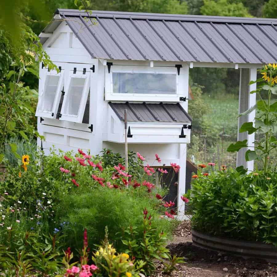 White chicken coop with a black roof surrounded by colorful flowers and greenery in a garden setting