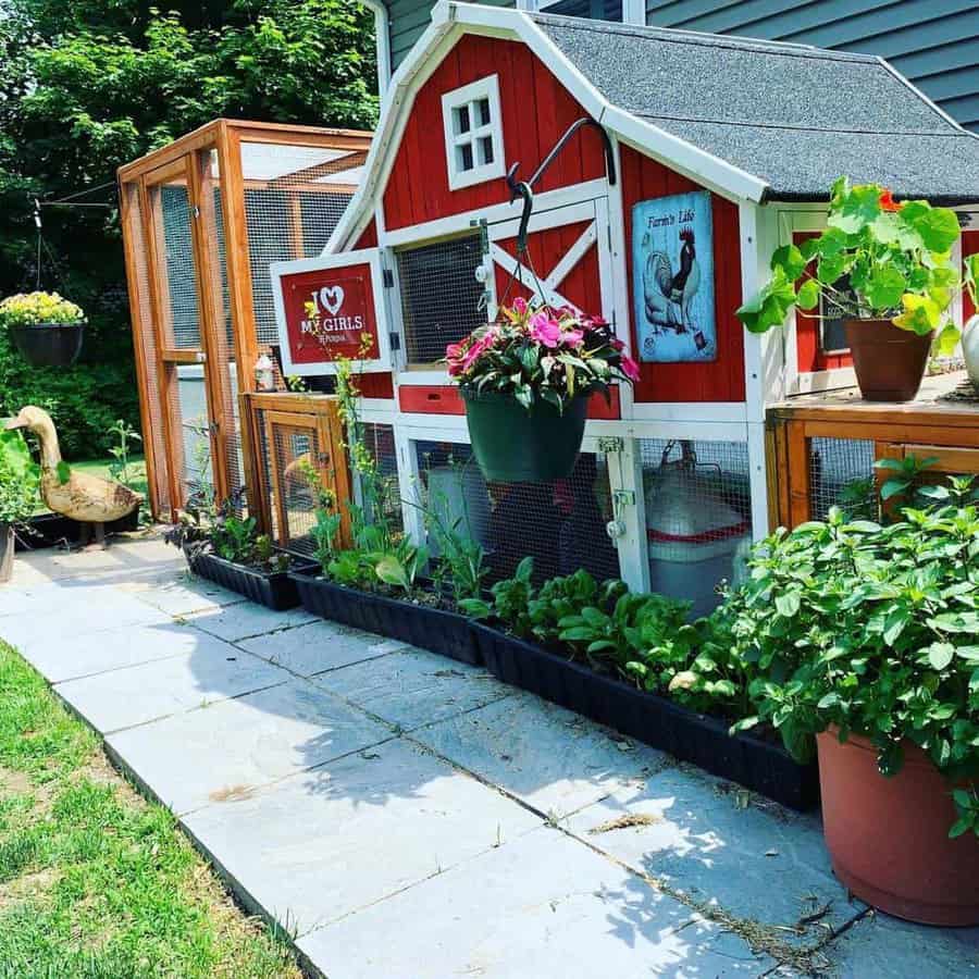 A red and white chicken coop with plants, a hanging flower pot, an "I ❤️ my girls" sign, and a goose statue on a stone path