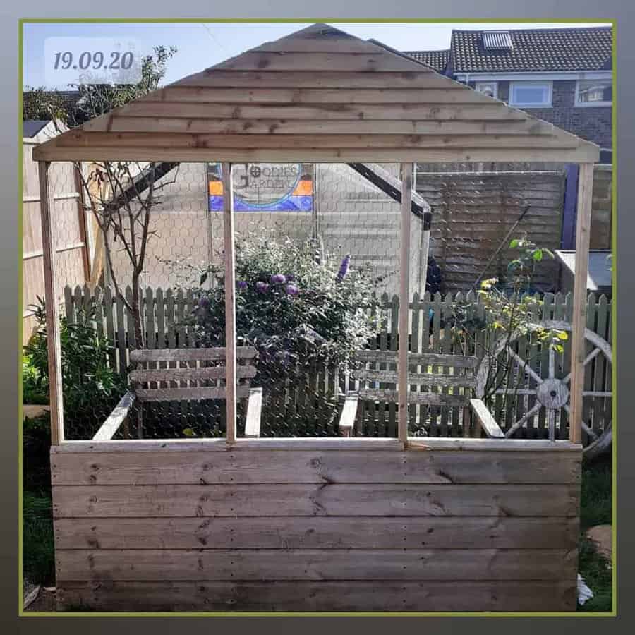 Wooden chicken coop with an open-air design, a slanted roof, and wire mesh, blending into a garden with flowers and fencing