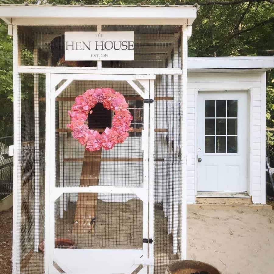 A white chicken coop labeled "The Hen House" with a pink wreath on the door, set in a green, wooded area