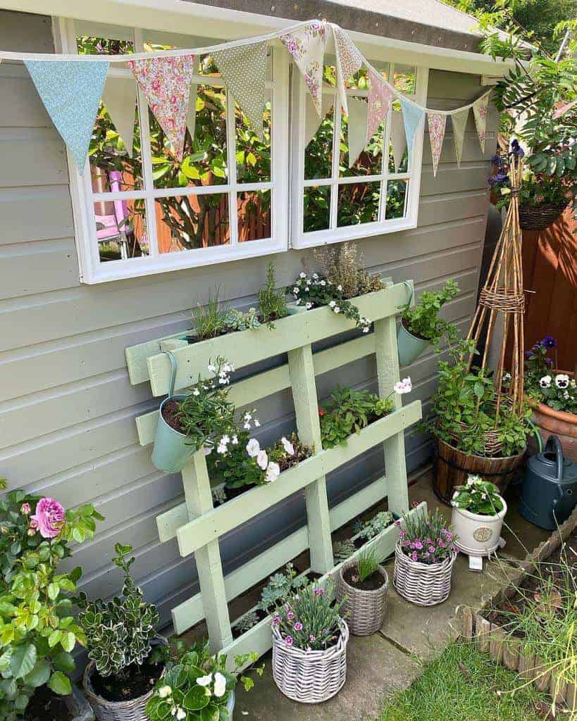 Charming pastel green pallet garden with blooming flowers, set against a cottage-style shed with bunting, wicker planters, and rustic decor