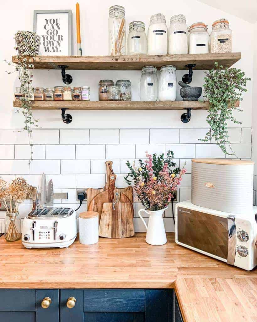 Bright kitchen with wooden shelves holding jars, spices, and plants; counter with a toaster, cutting boards, flowers, and a microwave