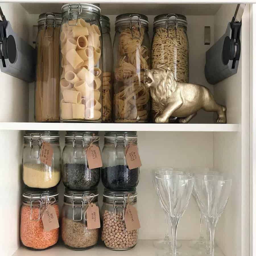 Pantry shelves with glass jars of pasta and grains, labeled tags, and a decorative lion figure; empty wine glasses below