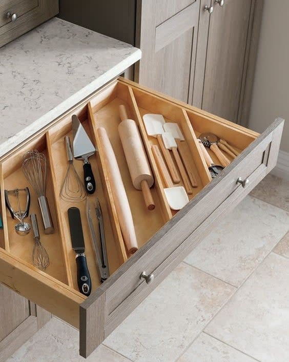 Open kitchen drawer with wooden dividers containing a rolling pin, spatula, whisk, pastry cutter, and measuring spoons