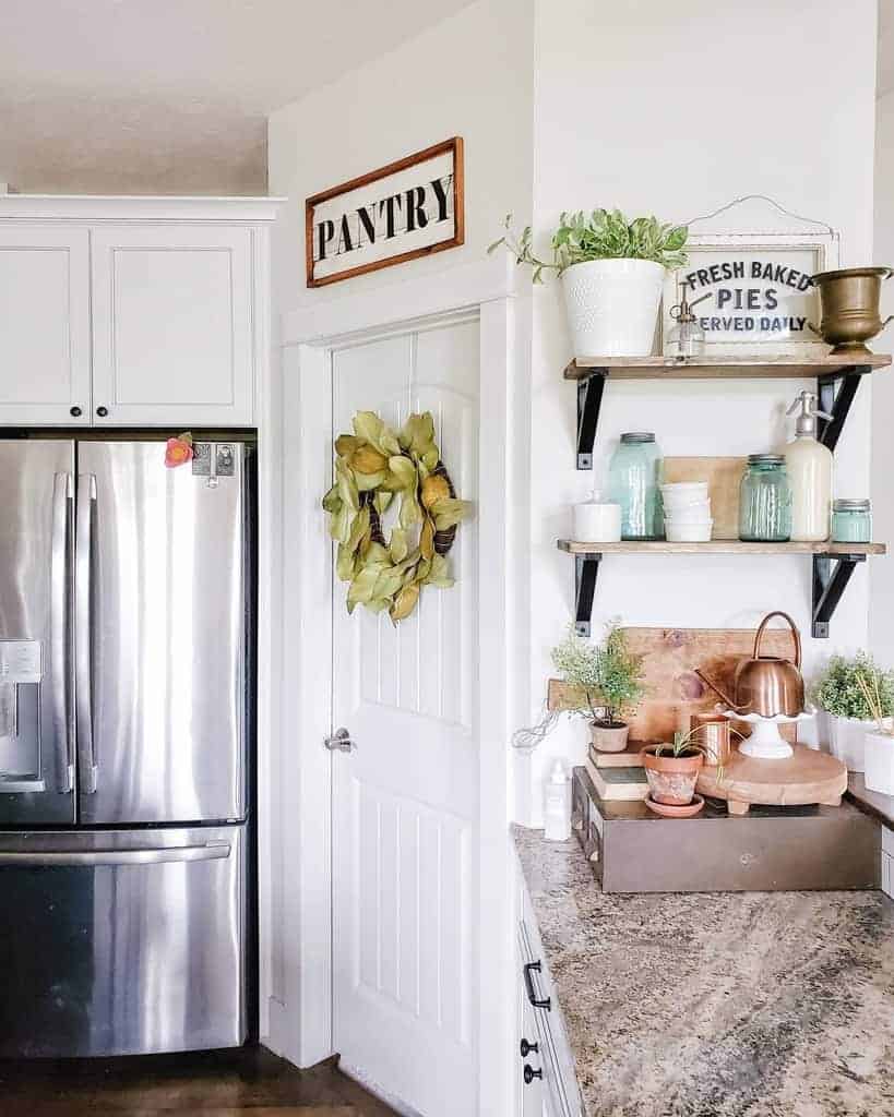 A cozy kitchen corner with stainless steel fridge, pantry door with wreath, and decorative shelves displaying plants and kitchenware