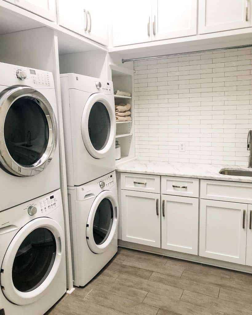 Modern laundry room with stacked washers and dryers, white cabinetry, built-in shelving, and a subway tile backsplash for a clean look