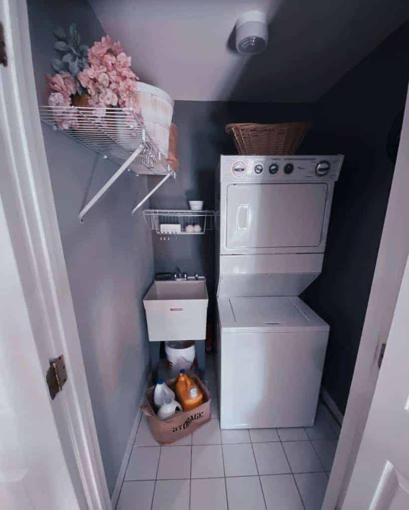 Compact laundry room with a stacked washer and dryer, small sink, shelves with flowers and supplies, and a laundry basket on top