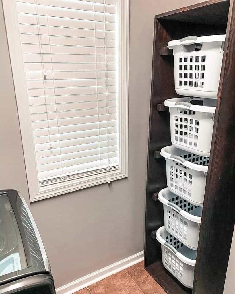 Five stacked white laundry baskets in a tall wooden shelf next to a window with white blinds in a laundry room