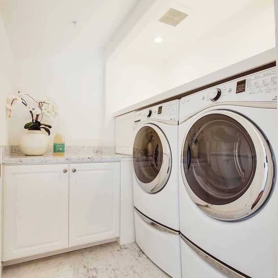 Bright laundry room with side-by-side front-loading washer and dryer, white cabinets, marble floor, and a potted orchid on the counter