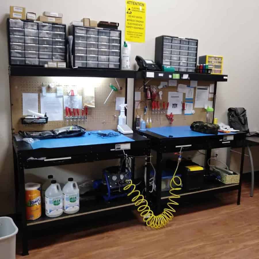 A workshop with two workbenches, organized tools, labeled boxes, cleaning supplies, and a yellow warning sign on the wall