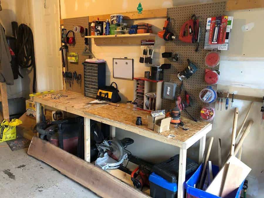 A cluttered workbench in a garage with tools, pegboard, shelves, and various equipment stored underneath