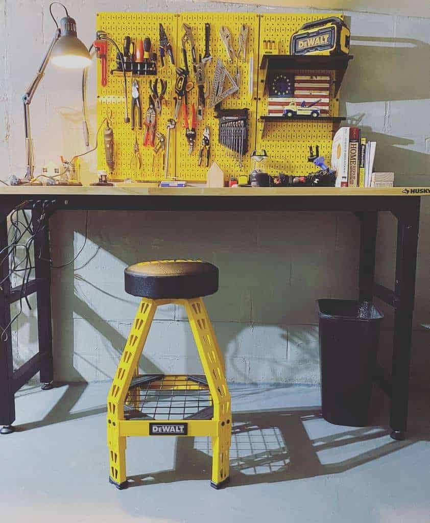 Workbench with tools hanging on a pegboard, a stool, a lamp, and a trash bin underneath marked with DeWalt brand