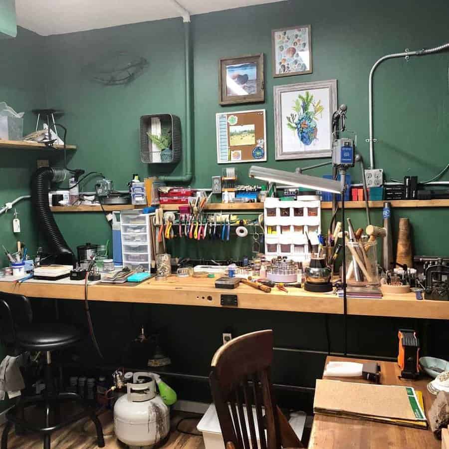 A cluttered workshop with tools, art supplies, framed pictures, and a wooden workbench against green walls