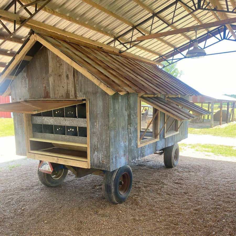 Mobile chicken coop on wheels with a corrugated metal roof and sides, under a shelter with a gravel floor