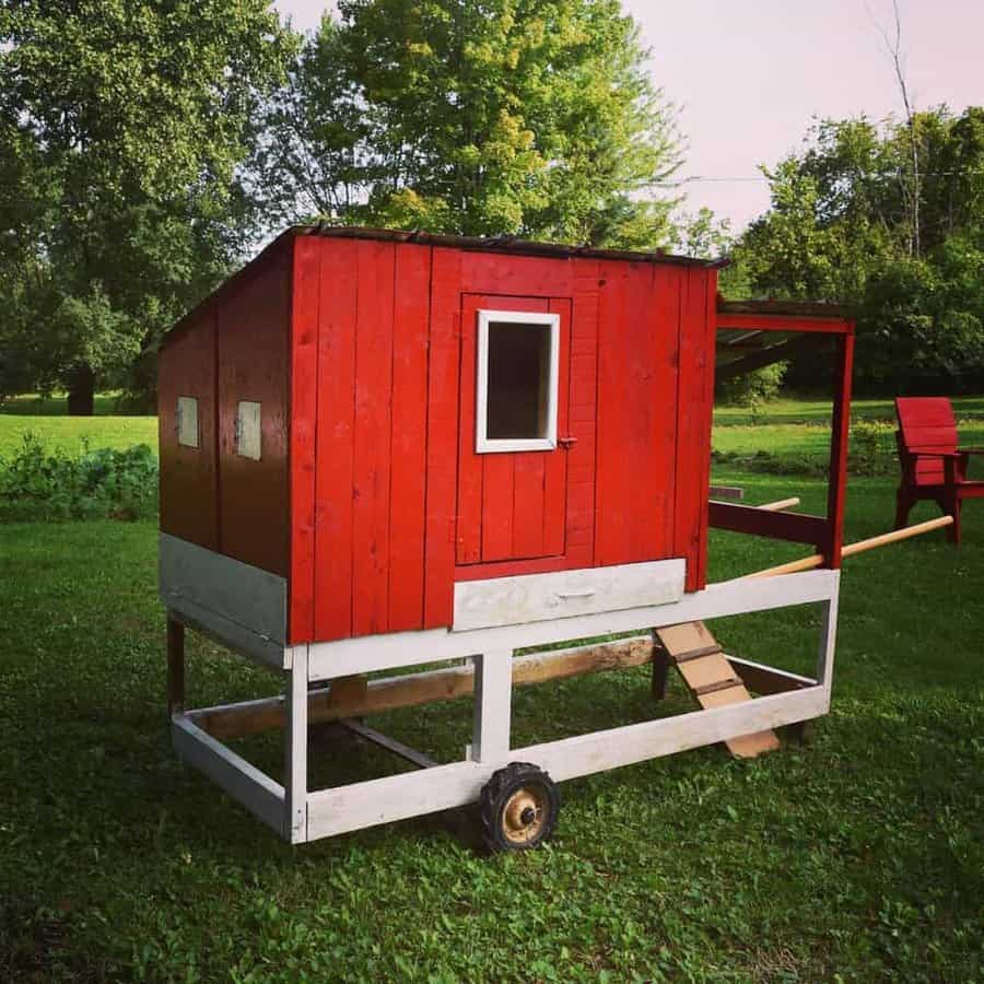 Red chicken coop on wheels in a grassy yard, surrounded by trees, with a small ramp leading to the entrance