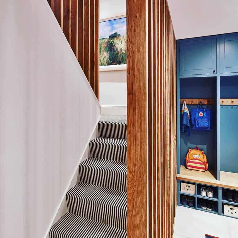 Staircase with striped carpet, wooden rails, and a hallway with blue cubbies containing backpacks and shoes