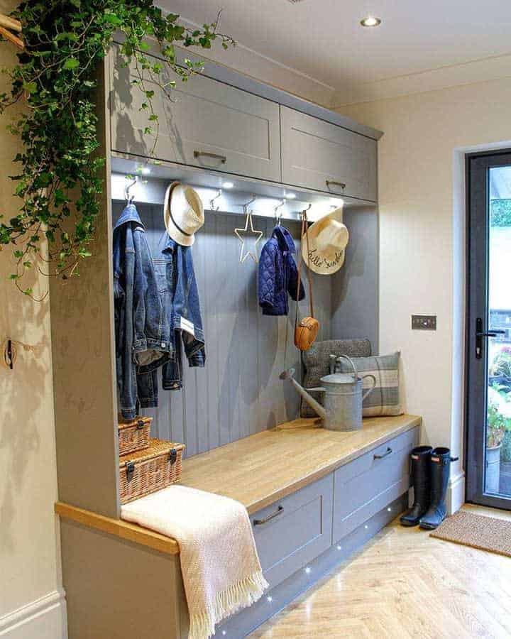 Mudroom with a wooden bench, jackets, hats, and a watering can. Above, greenery and decor hang, and a door on the right leads outside