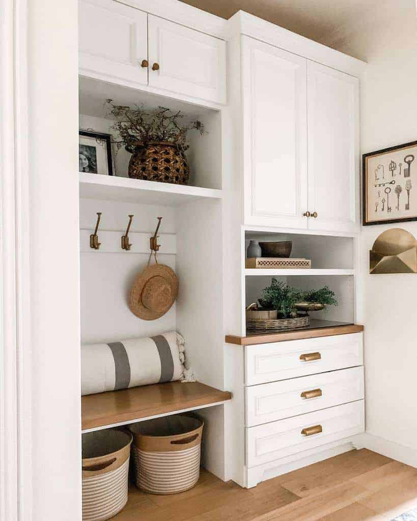 White entryway with built-in shelves, cabinets, hooks, and a bench; decor includes a basket, plants, a hat, and framed key artwork