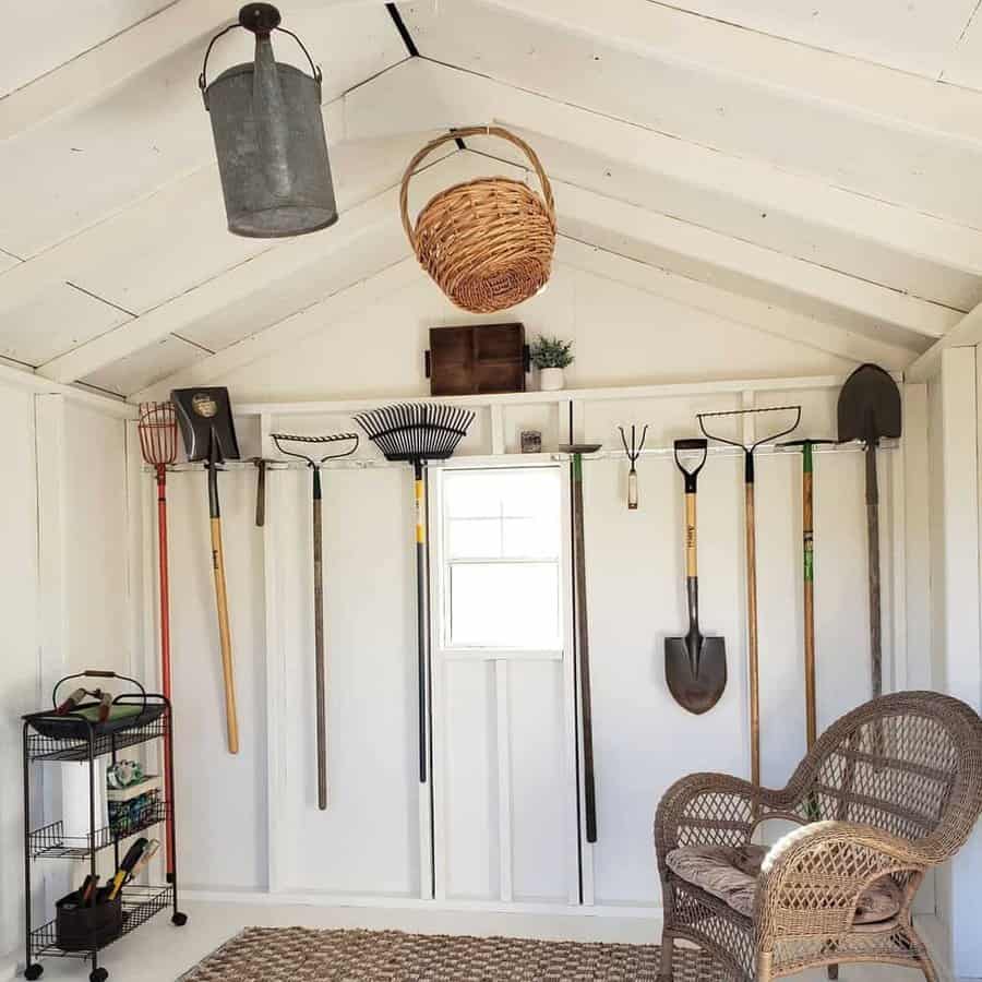 White shed with gardening tools on the wall, wicker chair, woven rug, metal bucket, basket, and a small cart in the corner