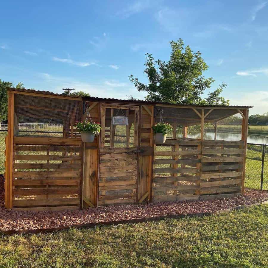 Rustic pallet wood chicken coop with a corrugated roof, mesh windows, and hanging planters, set in a scenic farm landscape