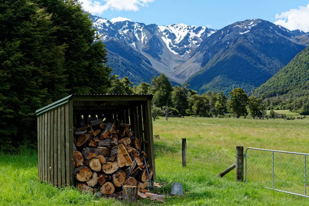 A woodpile in a shed on a grassy field with a fence, trees, and snow-capped mountains under a blue sky in the background