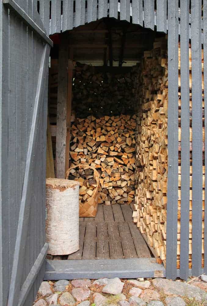 A wood shed with stacked firewood inside, partially open door, and a log with an axe resting on it