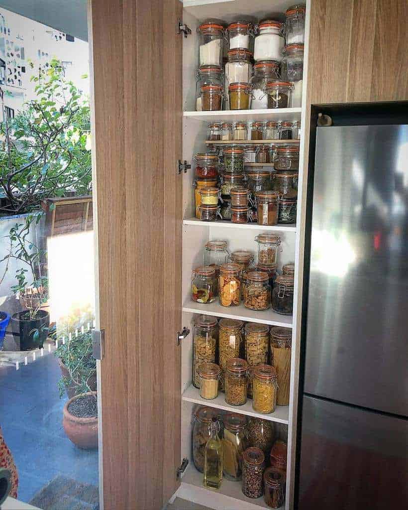 A pantry with wooden doors reveals shelves filled with jars of spices, grains, and pasta, next to a stainless steel refrigerator