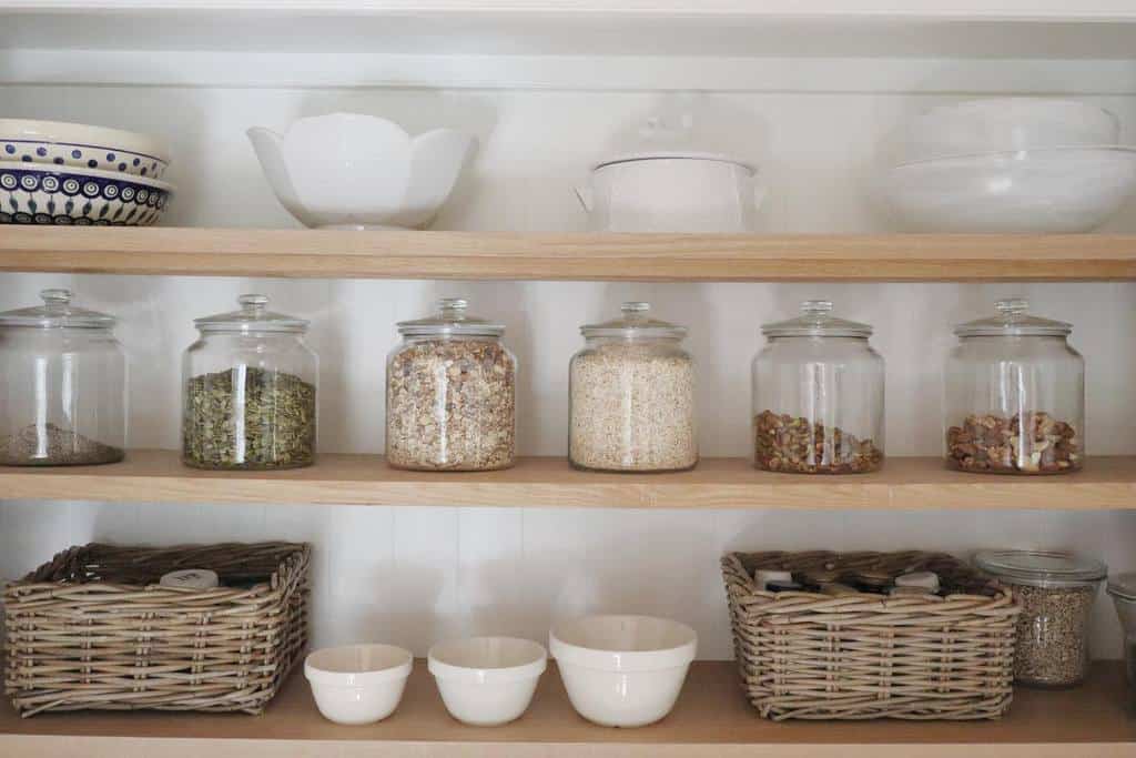 Pantry shelves with glass jars of grains, two wicker baskets, white bowls, and ceramic dishes displayed neatly