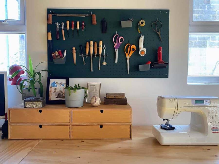 Sewing machine on a wooden table with a pegboard holding tools, scissors, and plants on drawers nearby