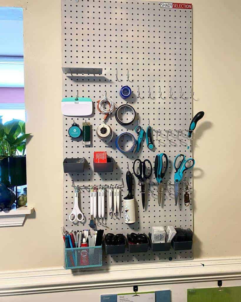 A pegboard on a wall holds tools like scissors, pliers, tape, and markers with a small plant on a shelf beside it