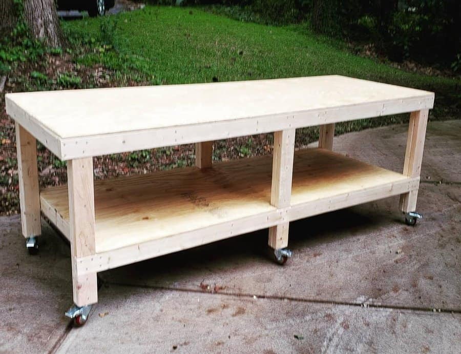 Wooden workbench on casters with a lower shelf, placed on a paved outdoor area, surrounded by grass and trees