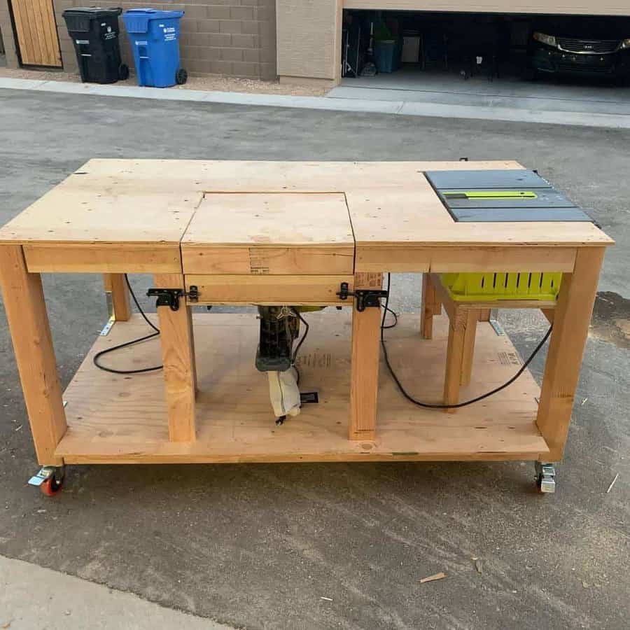 Wooden workbench on wheels with a saw installed, set outdoors near a garage and recycling bins