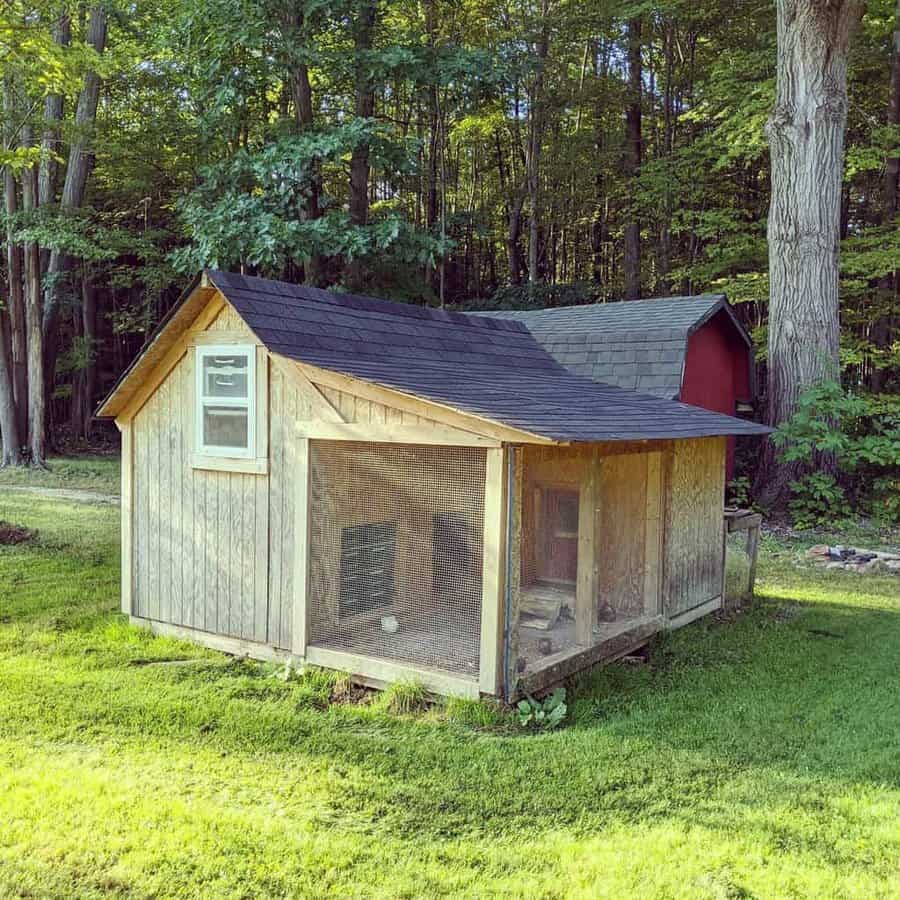 A small wooden chicken coop with a fenced area sits on green grass, surrounded by trees