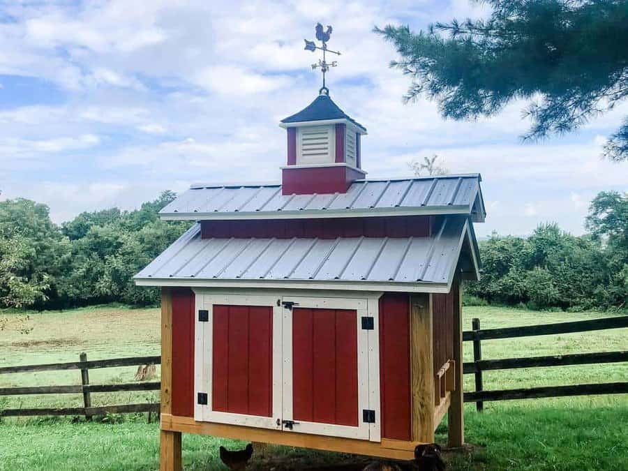 Red and white chicken coop with a weather vane on top, set in a green field with a wooden fence and trees in the background