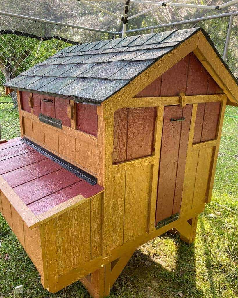 A small, colorful wooden chicken coop with a shingled roof and openable compartments, placed on a grassy area