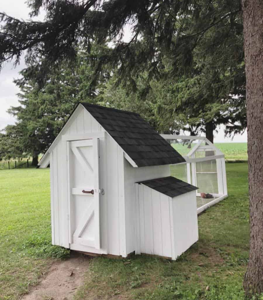 A small white chicken coop with a black roof, set in a grassy yard with surrounding trees