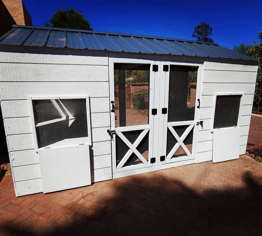 White wooden chicken coop with Dutch doors and mesh windows, featuring a metal roof for durability and ventilation