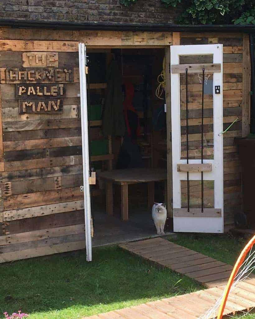 Rustic shed made from reclaimed wooden pallets, featuring a creative sign reading 'The Hackney Pallet Man' and a repurposed white door with metal accents