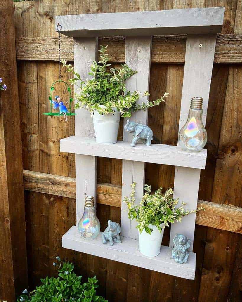 Charming upcycled pallet shelf painted in soft gray, mounted on a wooden fence, featuring potted plants, figurines, and decorative bulbs