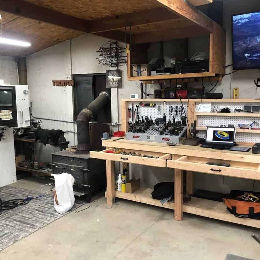 Woodworking shop with workbenches, tools, a stove with chimney, shelves, and a computer on a bench, wood ceiling beams visible