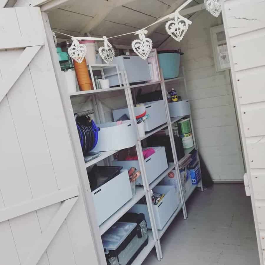 Open shed with organized shelves holding bins, paint cans, and tools. Decorative heart garland hangs above