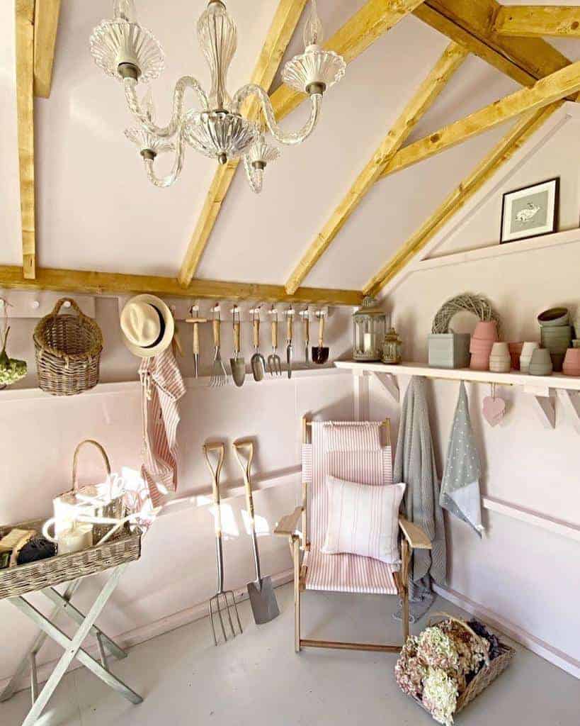 A cozy garden shed with tools on the wall, potted plants, a wicker chair and a chandelier, featuring wooden beams and soft pink walls