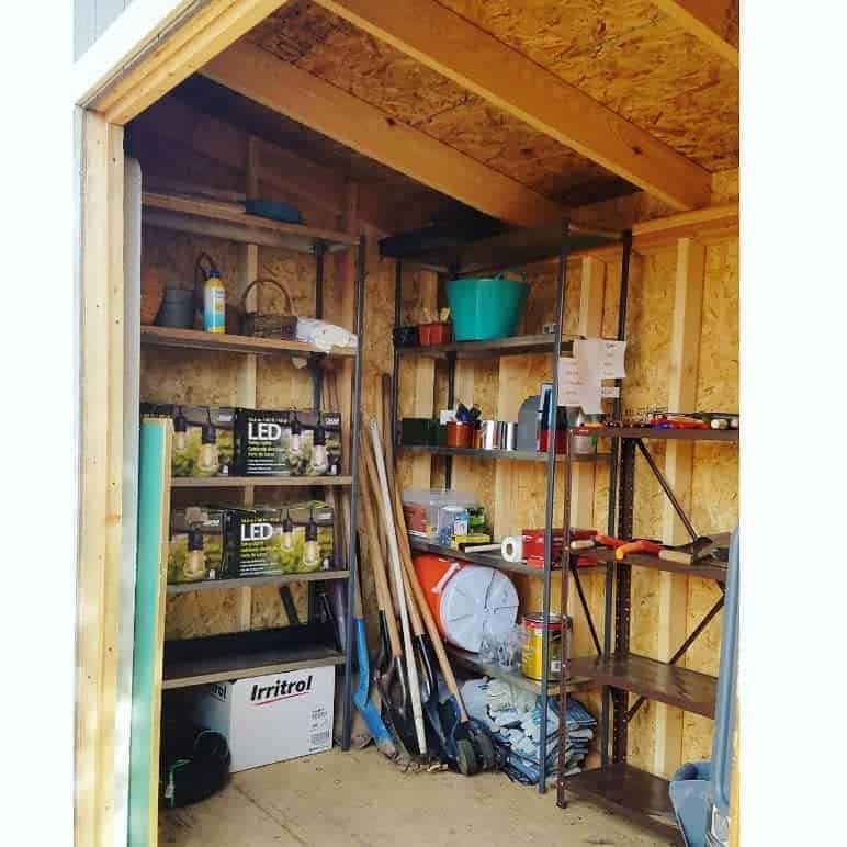 Inside a shed with shelves holding gardening tools, LED bulbs, a bucket, and various supplies; rakes and shovels are leaned against a wall