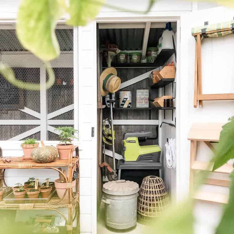 Small shed with gardening tools, straw hat, paint cans, and shelves; nearby, potted plants on a wooden table framed by green leaves