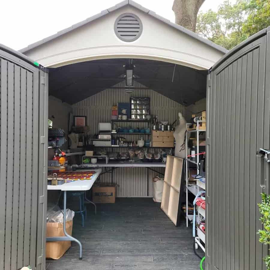 Open shed with shelves filled with tools and supplies, a mannequin on the right, tables holding various items, and trees visible outside