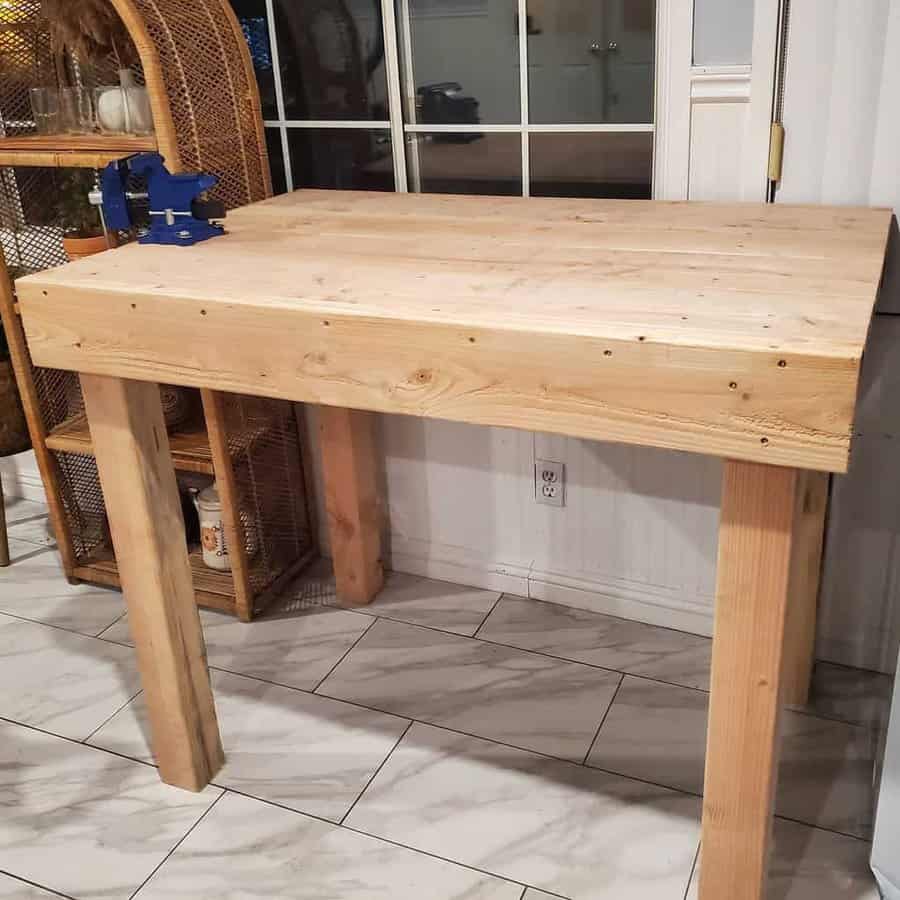 Wooden workbench with a blue vise on the corner, placed in a room with tiled floor and wicker shelving in the background