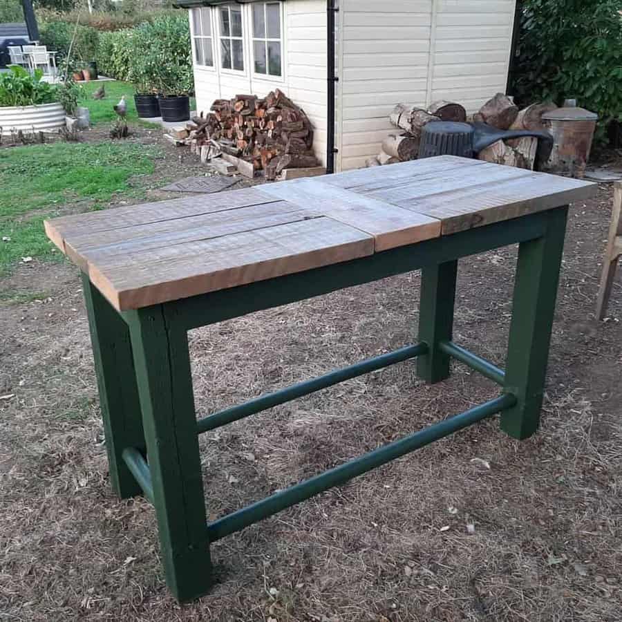 A rustic wooden table with a green frame stands outdoors on dry grass, with a shed and stacked firewood in the background