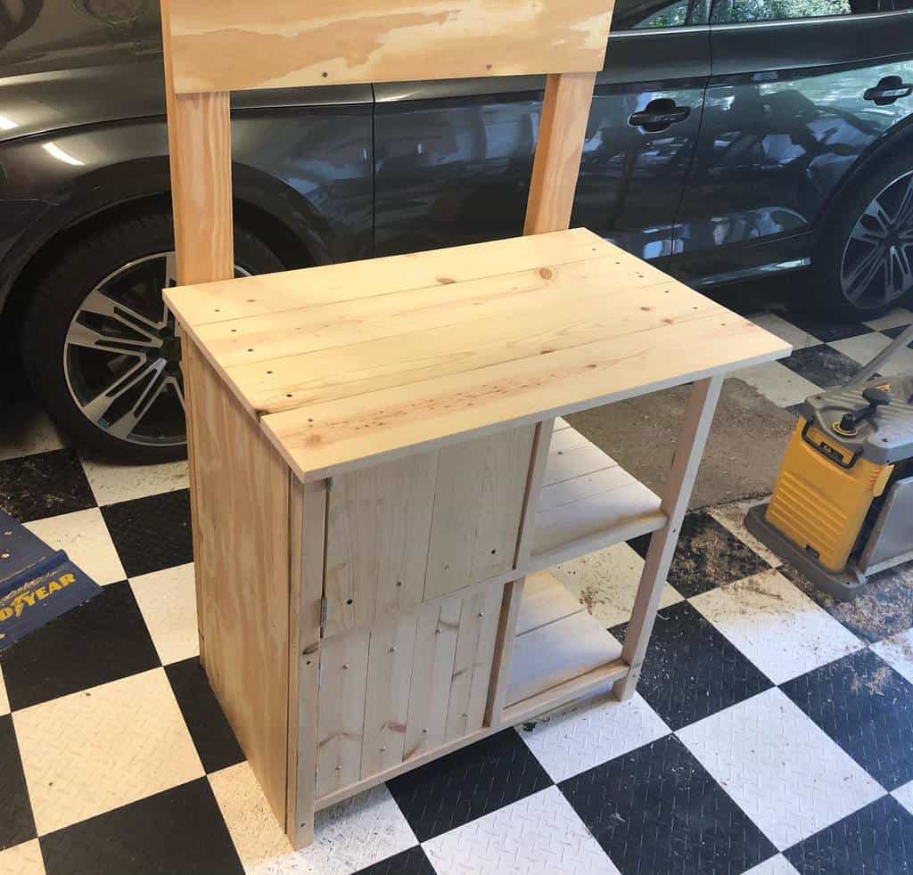 A wooden DIY workbench with shelves in a garage, on a black and white checkered floor next to a parked car