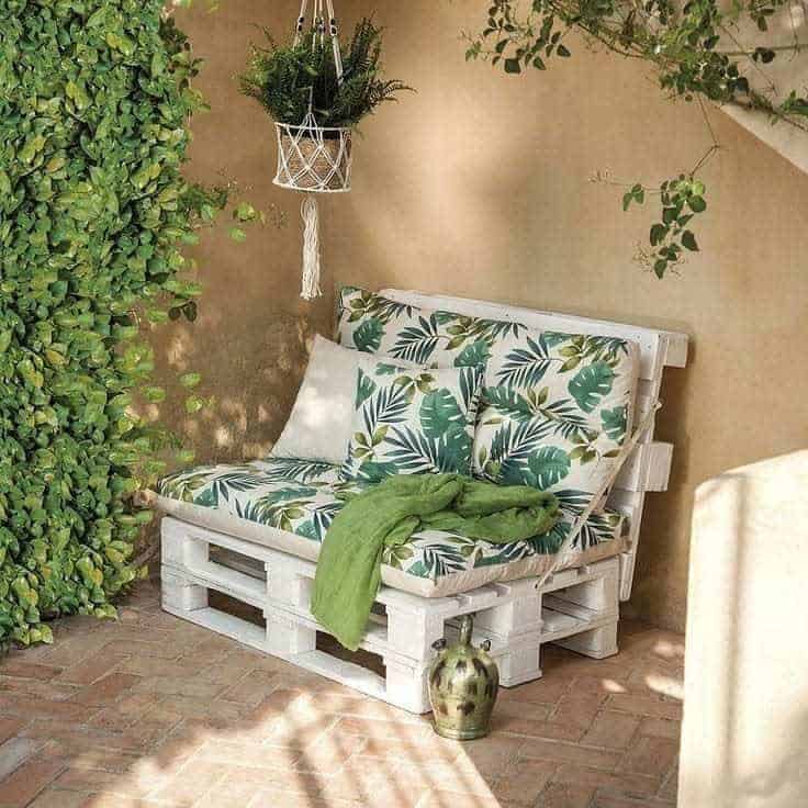 Cozy outdoor pallet sofa with white-painted wood, tropical leaf cushions, a green throw, and a hanging macramé plant holder